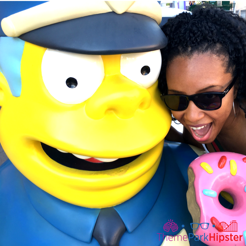 NikkyJ next to cop eating big pink doughnut Simpsons Springfield universal studios cop and doughnut with NikkyJ. Keep reading to learn about the cheap, best food at Universal Studios Orlando, Florida. #UniversalStudios #themepark #universalorlando with NikkyJ
