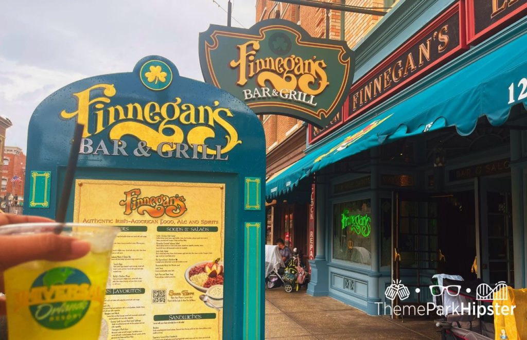 Universal Orlando Finnegan's Bar & Grill sign to present New York style Irish American and outdoor menu with colors of yellow and blue with ThemeParkHipster holding a drink up in the air. Keep reading to discover what are the best lounges at Universal Orlando.