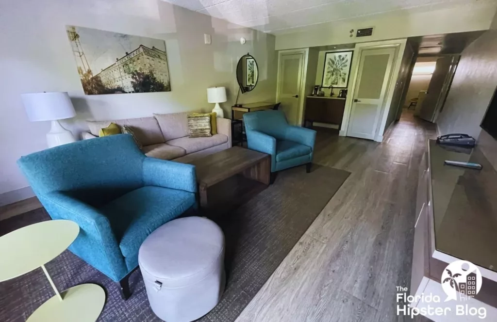 Hotel Alba Tampa Suite Room with cool tone colors of blues, brown, gray, and white featuring table and sofa and sofa chair seating in front of a mounted television. Keep reading to discover what are the best hotels near Busch Gardens Tampa.