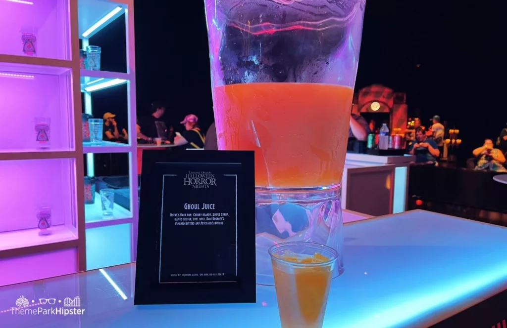 A cup of Ghoul Juice with a sign of ingredients in front of a big pitcher of Ghoul Juice at Universal Orlando Resort Halloween Horror Nights. Keep reading to discover more about Halloween at Universal. 