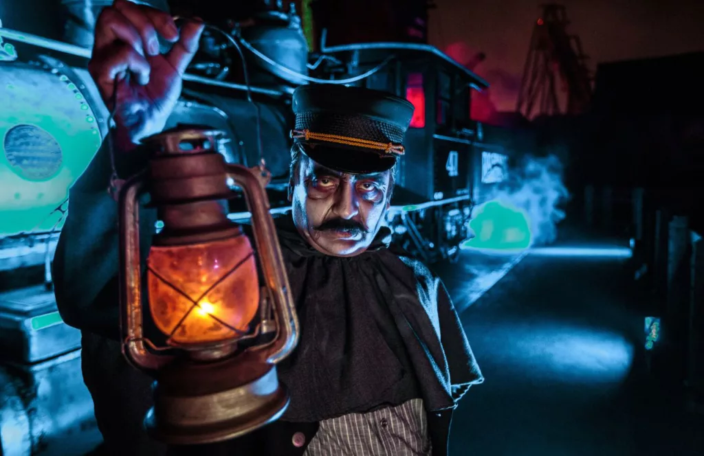 Ghost Town Scare Zone with a haunted train conductor holding an old lantern dressed from years past with a train and steam behind him at 2023 Knott's Scary Farm at Knott's Berry Farm in California. Keep reading to learn more about Knott’s Scary Farm houses.