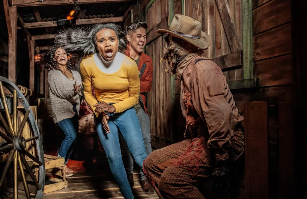 2023 Knott's Scary Farm at Knott's Berry Farm in California Origins_ The Curse of Calico House scene of people screaming and running scared through an old barn and a zombie in western wear. Keep reading to find out all you need to know about Knott’s Scary Farm houses.