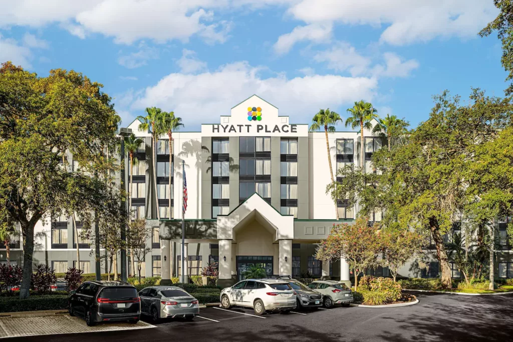 Multi-level Hyatt Place Tampa Busch Gardens Entrance with white exterior and black framed balconies with cars parked in the front parking lot surrounded by trees. Keep reading to discover what are the best hotels near Busch Gardens Tampa.