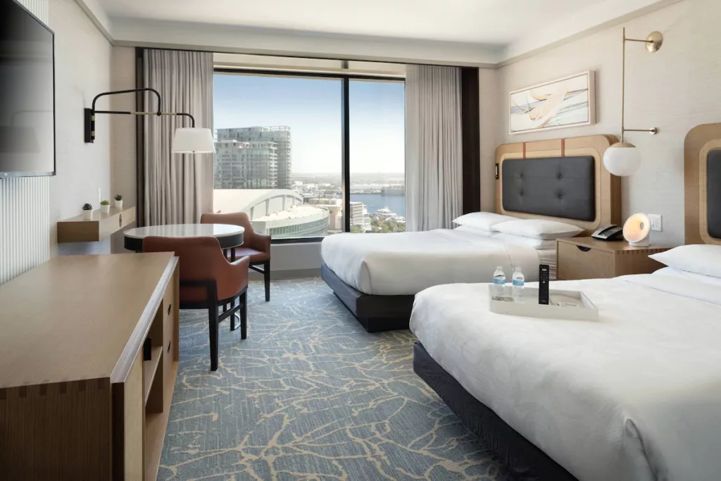 JW Marriott Tampa Water Street Standard Room overlooking the River and Tampa skyline with seating area at the window, mounted television above the desk and two large beds with amenities on the bed. Keep reading to discover what are the best hotels near Busch Gardens Tampa.