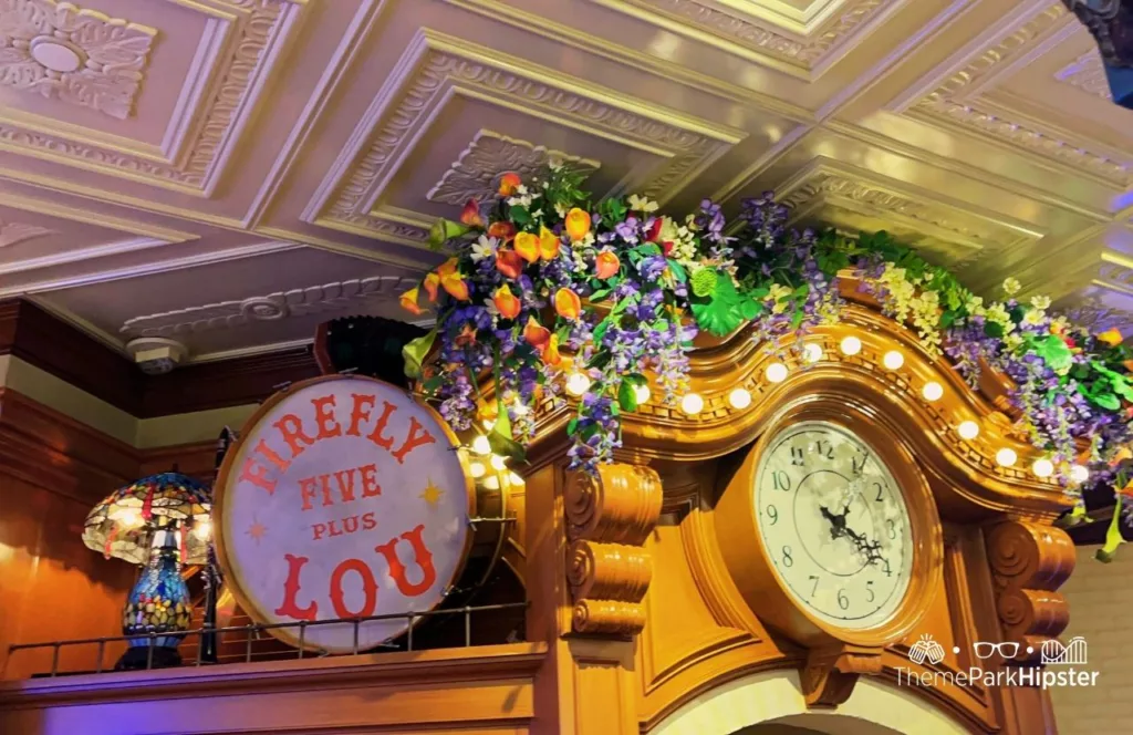 2024 Floral arrangement on the grand clock and a vintage drum of display that says Firefly Five Plus Lout at Princess and the Frog Tiana's Palace Restaurant at Disneyland. Keep reading to discover more about Tiana’s Palace food.