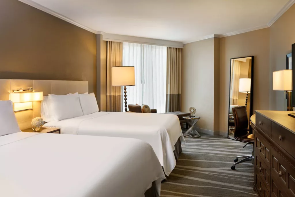 Renaissance Tampa International Plaza Hotel Standard Room with ambient lighting, crisp white bedding, large window with seating as well as a mirror and grand dresser.Keep reading to learn more about the best hotels near Busch Gardens Tampa.
