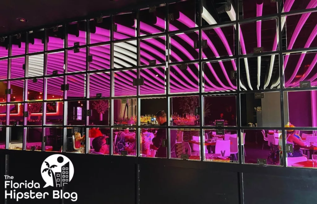 View of STK Orlando from the exterior looking in to a wall of windows with purple and white lighting inside and black building exterior. Keep reading to find out all there is to know about STK Orlando. 