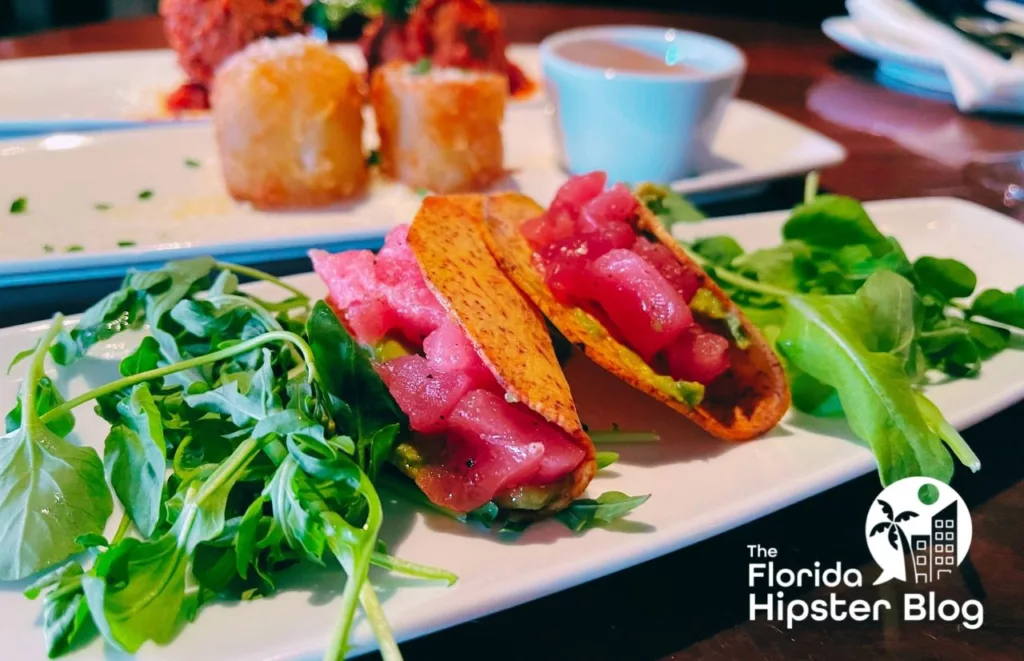 STK Orlando Tuna Poke served in a crispy tortilla taco alongside greens and tater tots the background. Keep reading to discover more about STK Disney Springs.