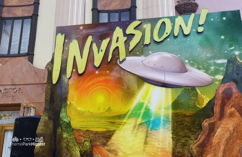 Invasion Sign with a UFO shining its lights down on the earth's surface at Lights, Camera, Hacktion Eddie's Revenge HHN 30 Scare Zone at Halloween Horror Nights 2021. Keep reading to discover more about Scare zones at Halloween Horror Nights.
