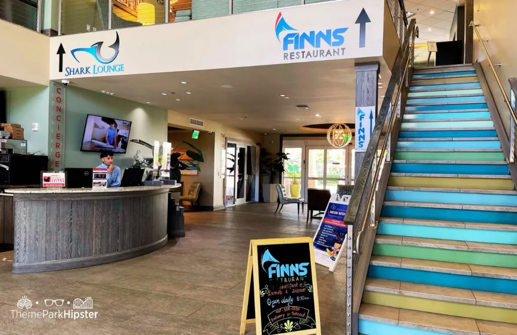Lobby area in Encore Resort with stairs leading up to Finns Restaurant, signage for Shark Lounge and concierge desk with staff. Keep reading find out all you need to know about Encore Resort in Orlando. 